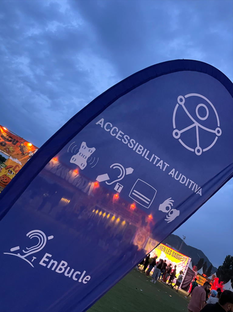 Bandera amb senyalització d’accessibilitat auditiva en un esdeveniment a l’aire lliure, amb icones de bucle magnètic, subtitulació i llengua de signes.