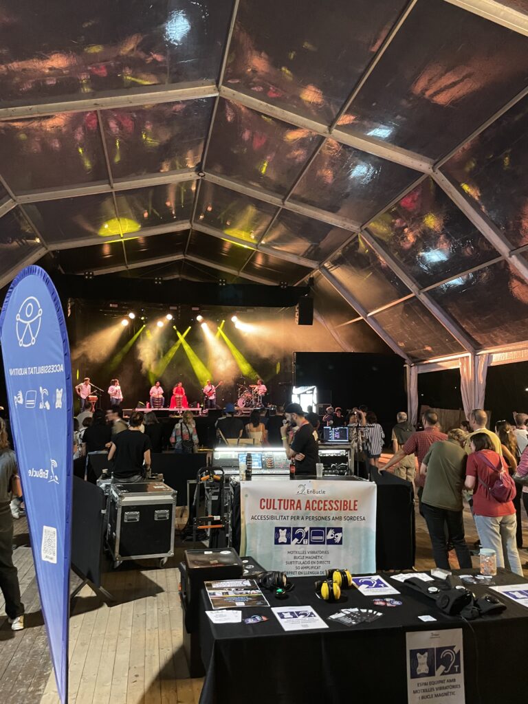 Espai d’accessibilitat amb taula informativa i equips com motxilles vibratòries, bucle magnètic i auriculars, davant d’un escenari amb música en directe i públic assistent.