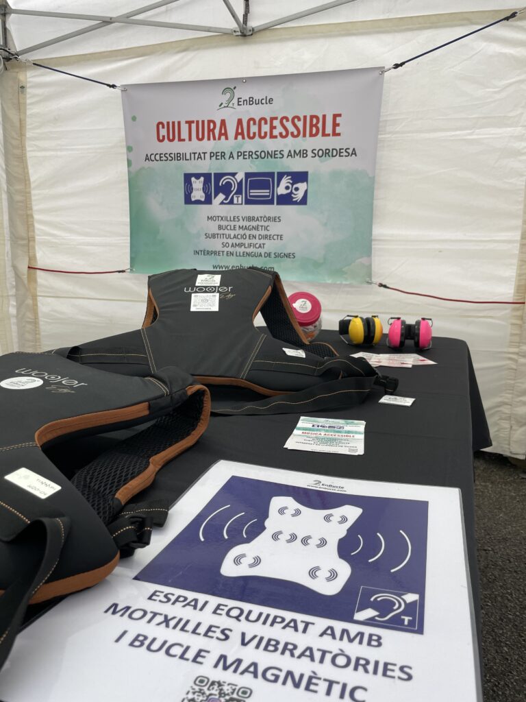 Taula amb equips d’accessibilitat auditiva en un estand, amb motxilles vibratòries, receptors i cartells informatius sobre cultura accessible.