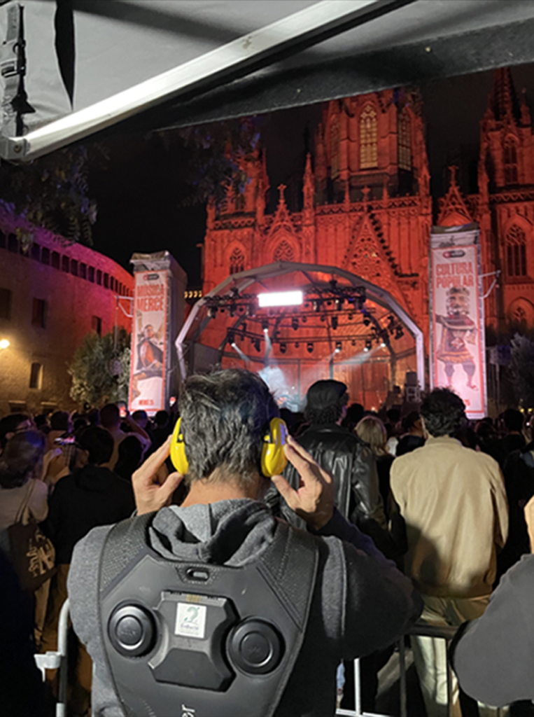 Persona amb motxilla vibratòria durant un concert a l’aire lliure, amb escenari il·luminat i públic al voltant.